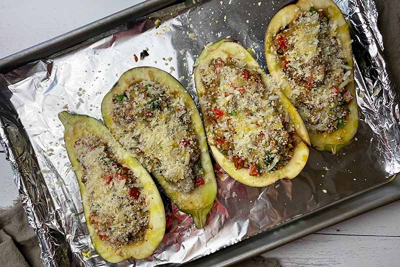 Horizontal image of unbaked vegetable halves with a filling and breadcrumb topping on a foil-lined sheet pan.