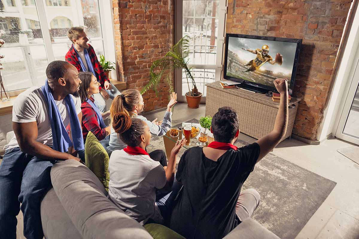 Horizontal image of an excited group of friends watching football at home while eating assorted food and drinking beer.
