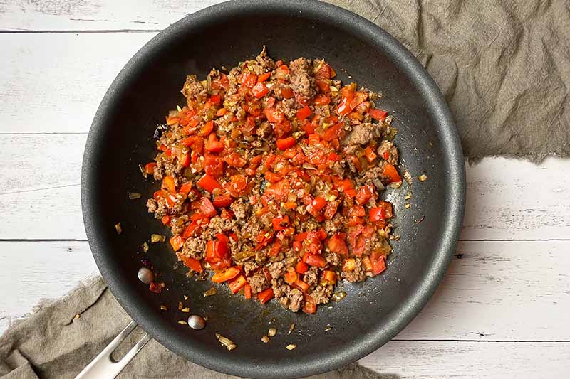 Horizontal image of cooking a ground meat, tomato, and bell pepper mix in a pan.