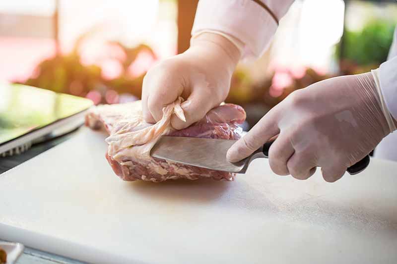Horizontal image of trimming the fat off raw meat with a knife while wearing gloves.
