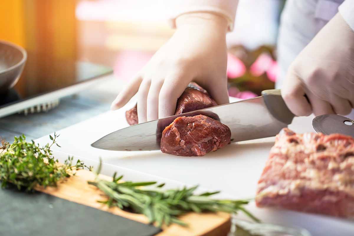Horizontal image of slicing into a raw piece of meat while wearing gloves in front of fresh herbs.