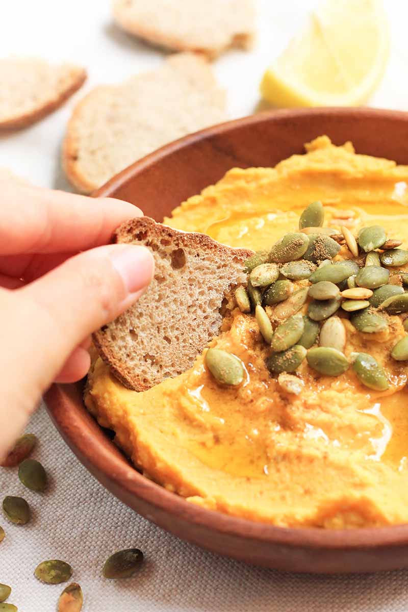 Vertical image of a hand scooping some bright orange hummus in a wooden bowl onto a thin slice of bread.