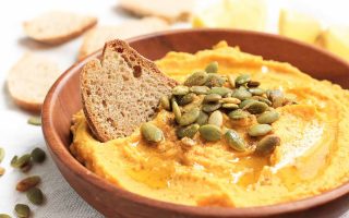 Horizontal image of a wooden bowl filled with a bright orange hummus topped with seeds with a slice of bread inserted into it.