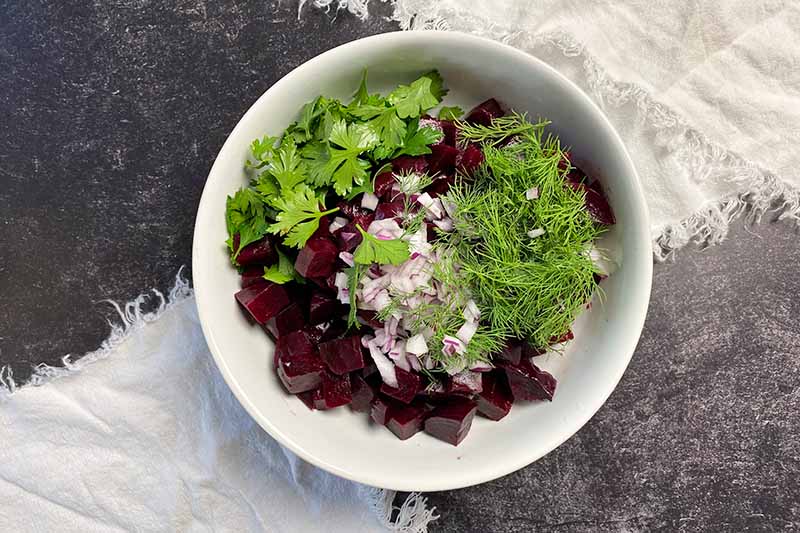 Horizontal image of piles of fresh herbs, red onion, and purple cubed vegetables in a large white bowl.