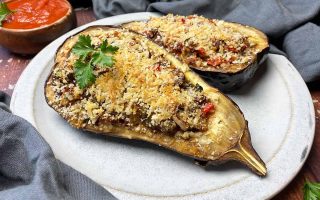 Horizontal image of two eggplant halves on a plate with a meat and mixed veggie filling and breadcrumb topping, next to gray napkins and a bowl of tomato sauce.