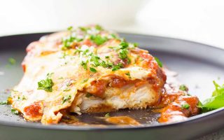 Horizontal image of a breaded poultry breast topped with tomato sauce and melted mozzarella cheese on a black plate with herb garnish.