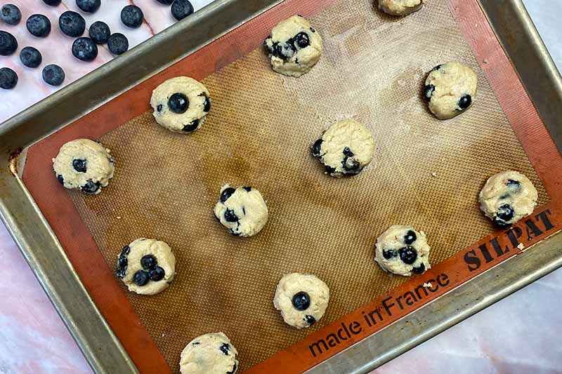 Horizontal image of unbaked mounds of dough topped with fresh fruit on a baking sheet lined with a silicone mat.
