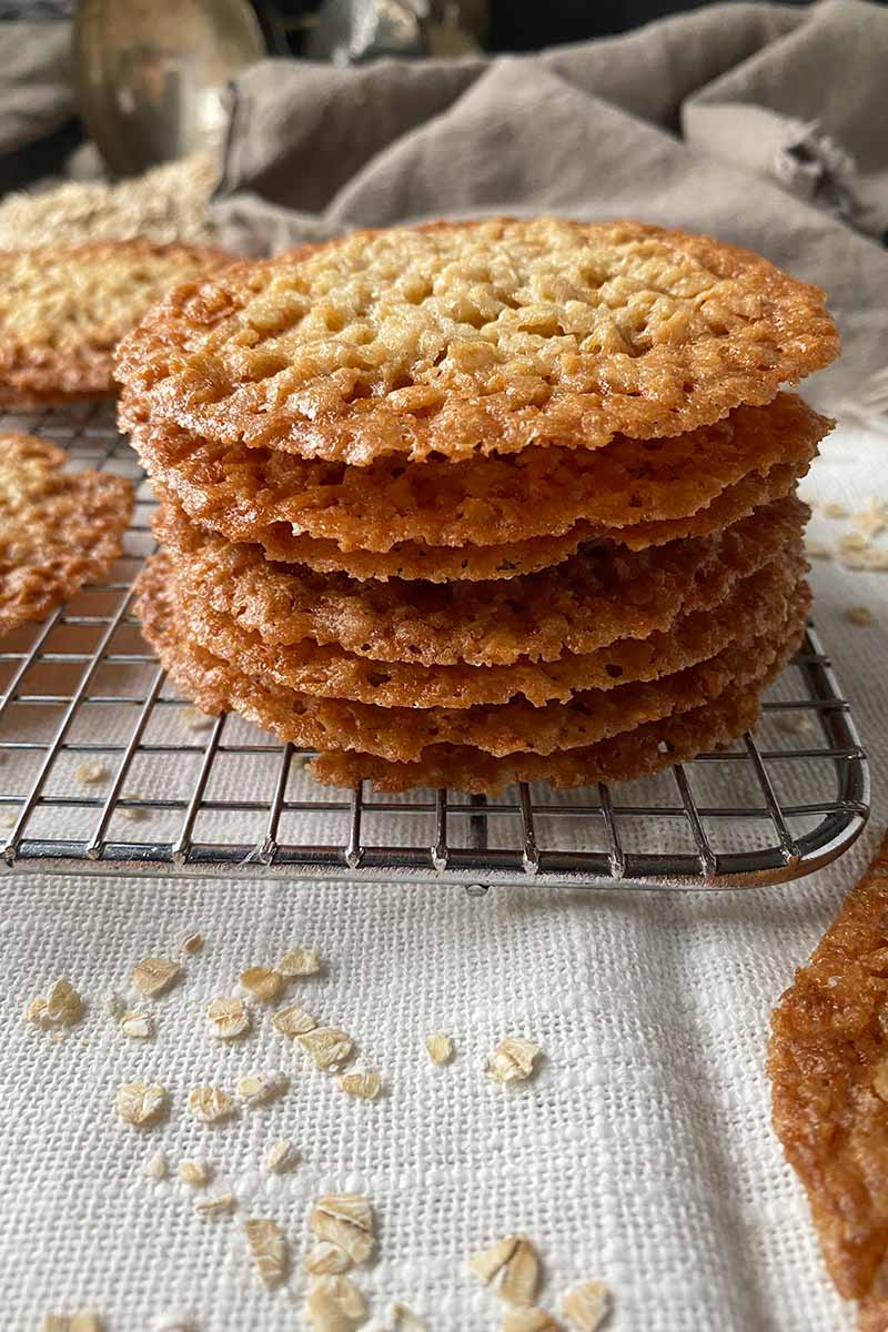 Vertical image of a stack of thin cookies on a cooling rack on a towel next to oats.