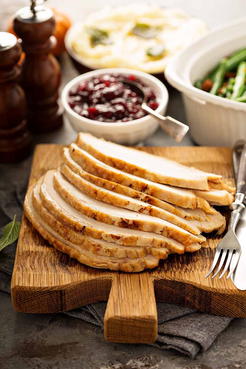 Vertical image of sliced turkey breast on a wooden board in front of dishes of cranberry sauce, mashed potatoes, green beans, and salt and pepper mills.