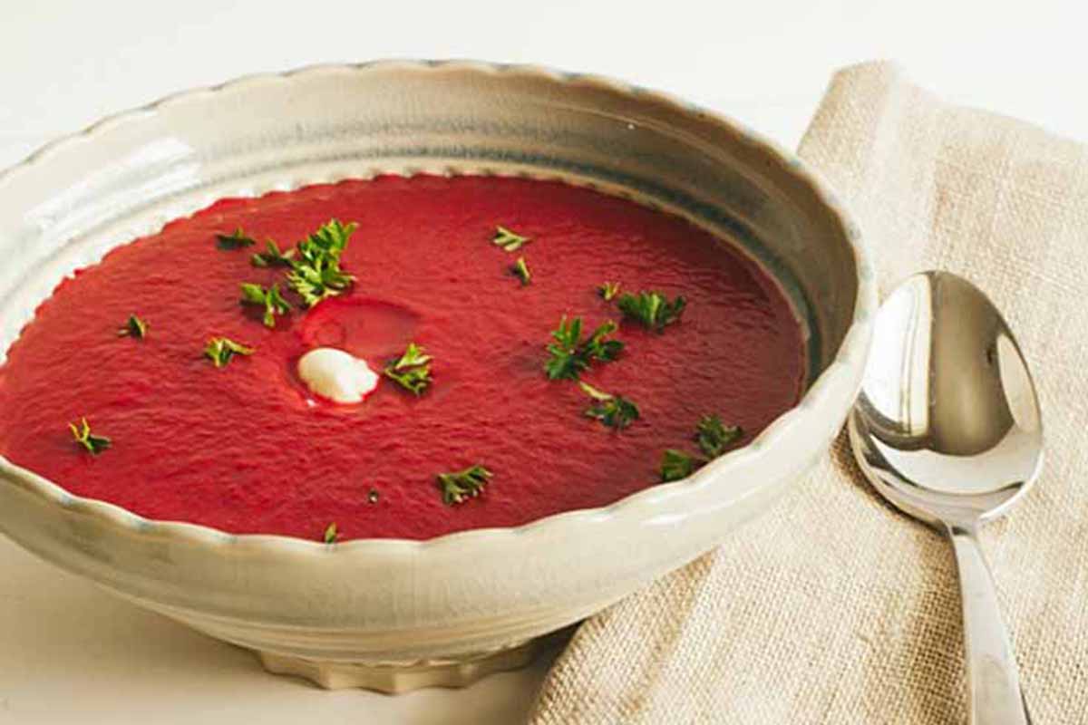 Horizontal image of creamy red soup in a bowl with herb garnish.