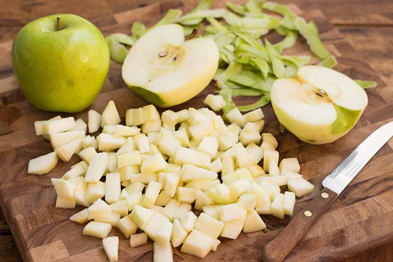 Horizontal image of diced and halved Granny Smiths on a wooden cutting board next to a serrated knife.
