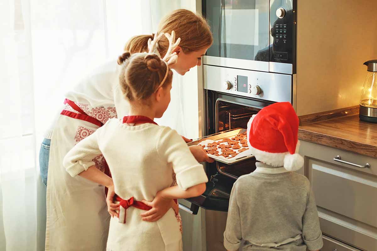 Horizontal image of a mom pulling gingerbread men out of the oven while a brother and sister watches.