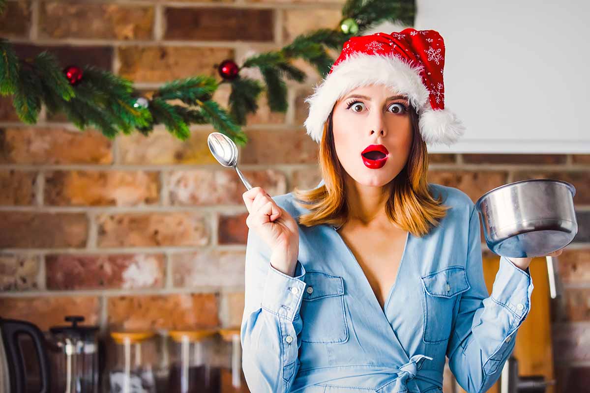 Horizontal image of a stressed woman in a Santa hat holding a pot and spoon.