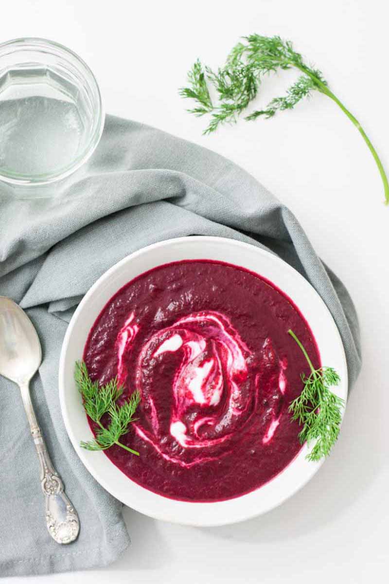 Vertical top-down image of a deep magenta soup with cream swirls on a blue napkin next to fresh dill.