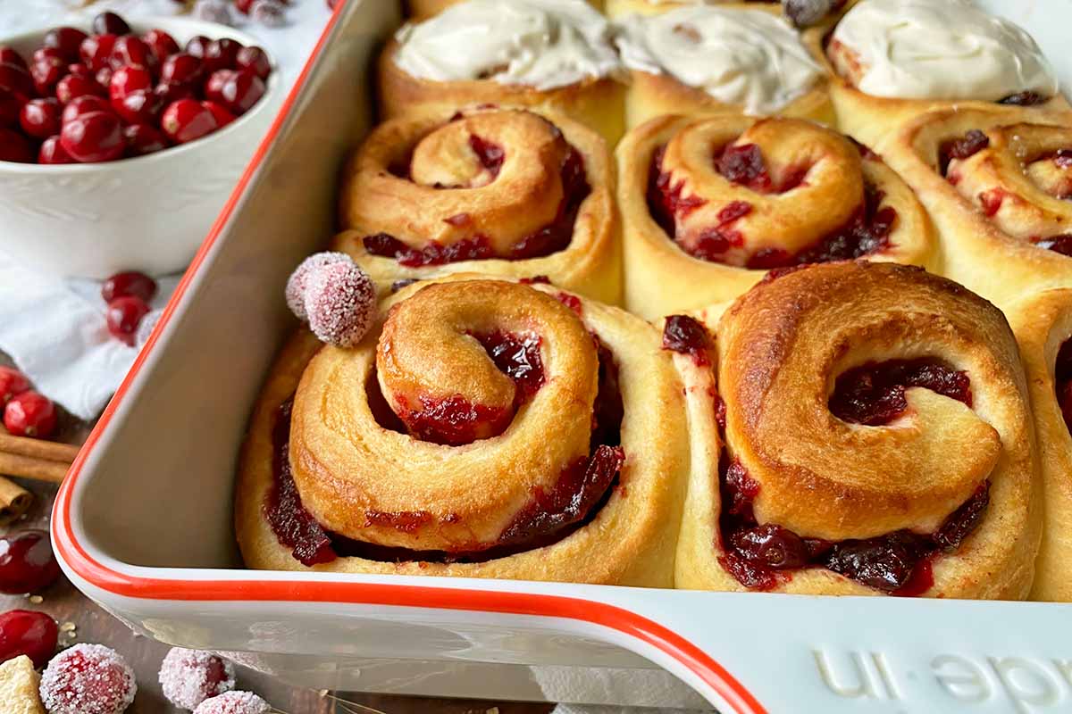 Horizontal close-up image of cranberry cinnamon rolls in a casserole dish, half of them frosted.