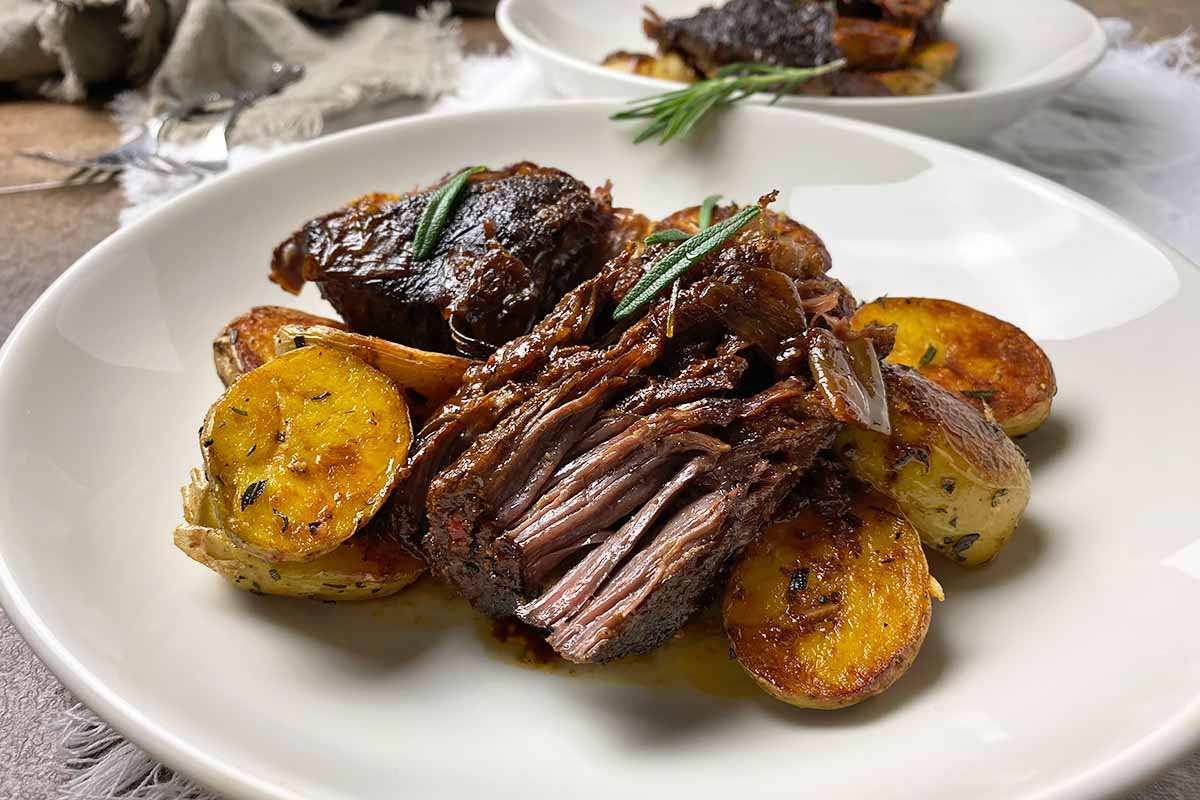 Horizontal image of a composed plate with chunks of beef on a bed of roasted potatoes with a rosemary herb garnish.