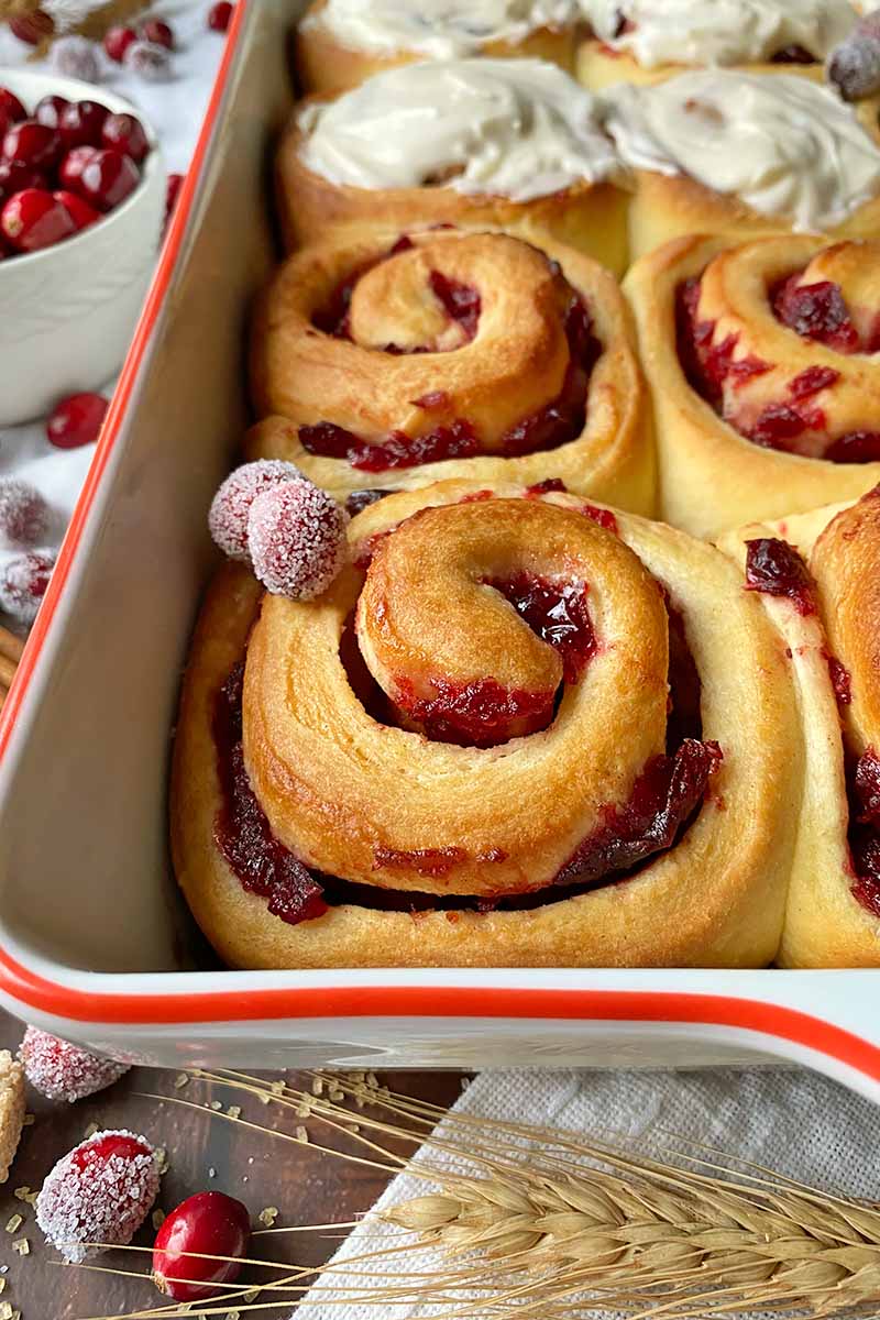 Vertical close-up image of rows of cinnamon rolls with a red fruit swirl, half of them frosted, in a casserole dish.