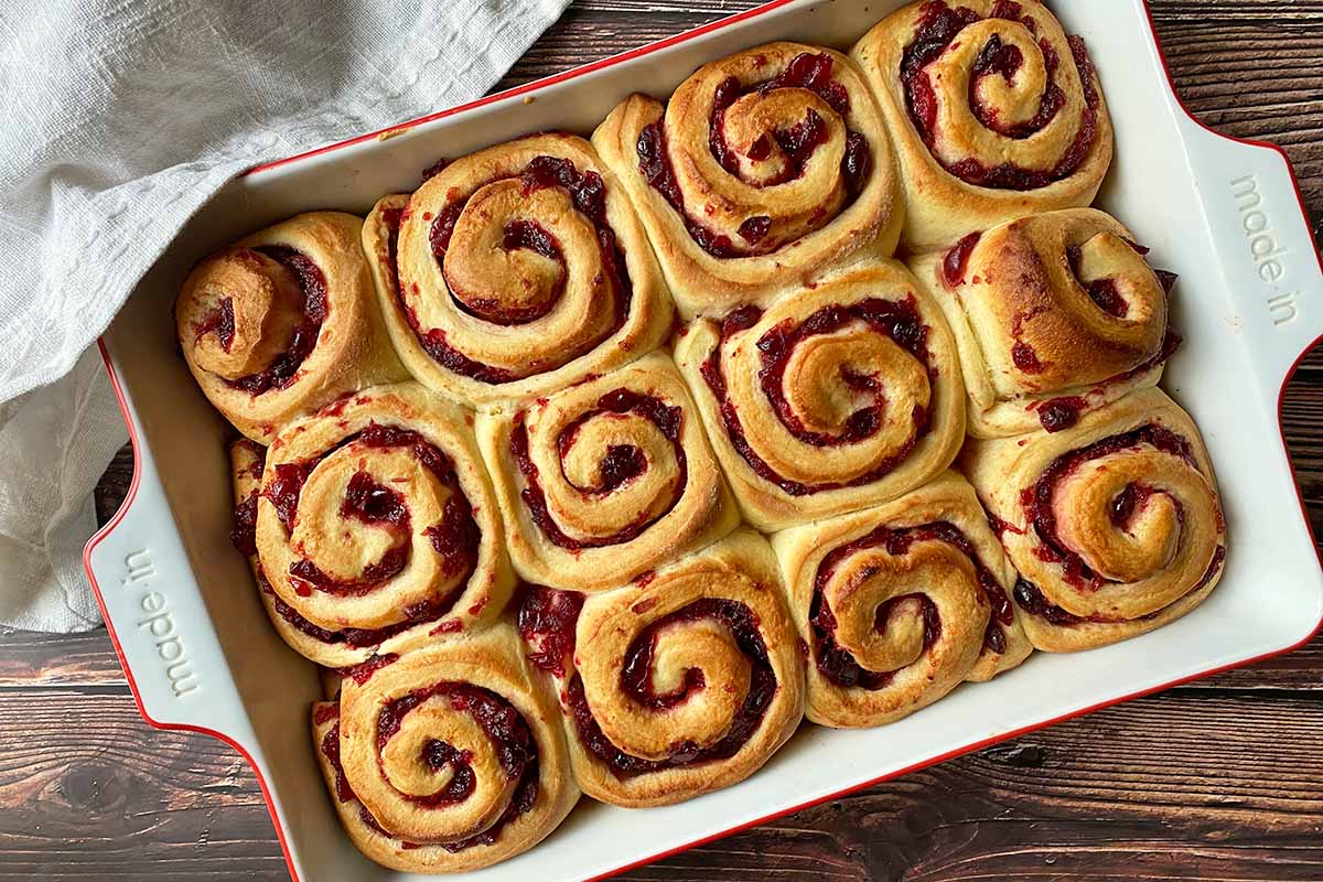 Horizontal image of freshly baked fluffy pastry pinwheels in a casserole dish with a cranberry sauce swirl.