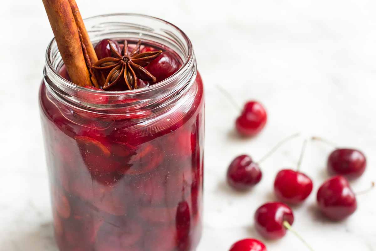 Horizontal image of a jar full of preserved cherries in syrup topped with a stick of cinnamon and star anise.