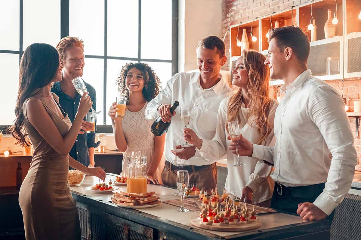 Horizontal image of a group of friends celebrating together with appetizers and drinks in the kitchen.