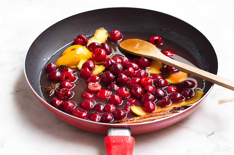 Horizontal image of heating a sugary syrup with red fruit, citrus peels, and spices in a skillet.