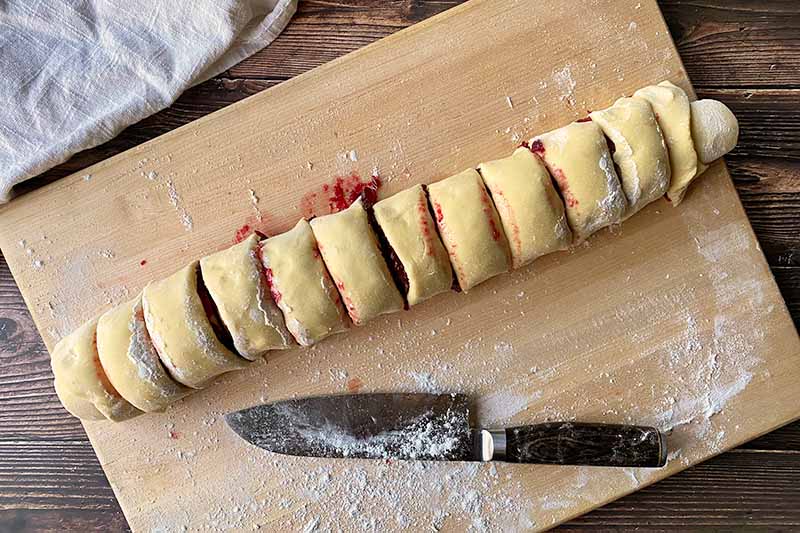 Horizontal image of a portioned log of dough with a red fruit filling on a lightly floured surface next to a dusted chef's knife.