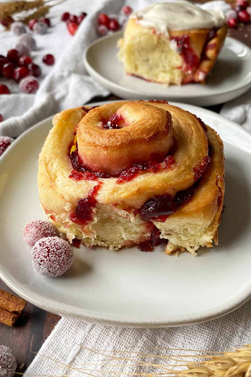 Vertical image of two cinnamon rolls with a cranberry swirl on white plates, one with cream cheese frosting.