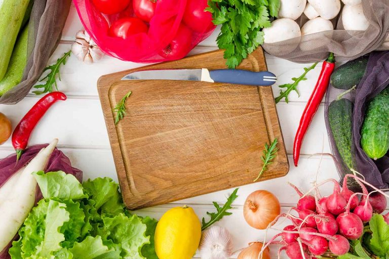 Horizontal image of a wooden cutting board and knife surrounded by assorted produce items.