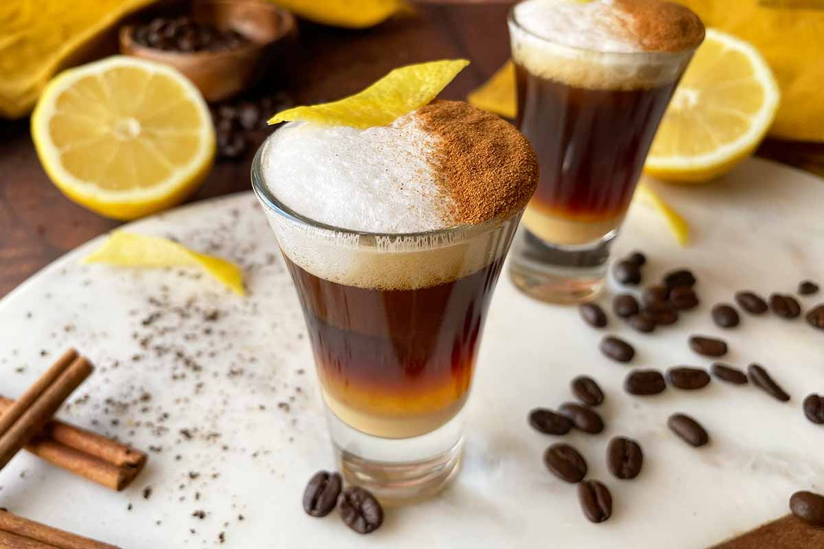 Horizontal image of two shooters filled with a layered espresso beverage topped with frothed milk, cinnamon, and lemon peel.