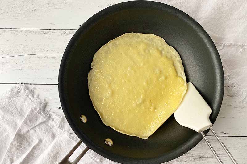 Horizontal image of preparing to flip a crepe with a rubber spatula in a nonstick skillet.