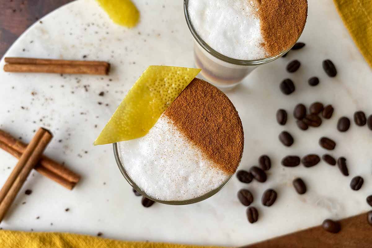 Horizontal image of two shooters filled with an espresso drink topped with frothed milk and garnished with cinnamon and lemon peel.