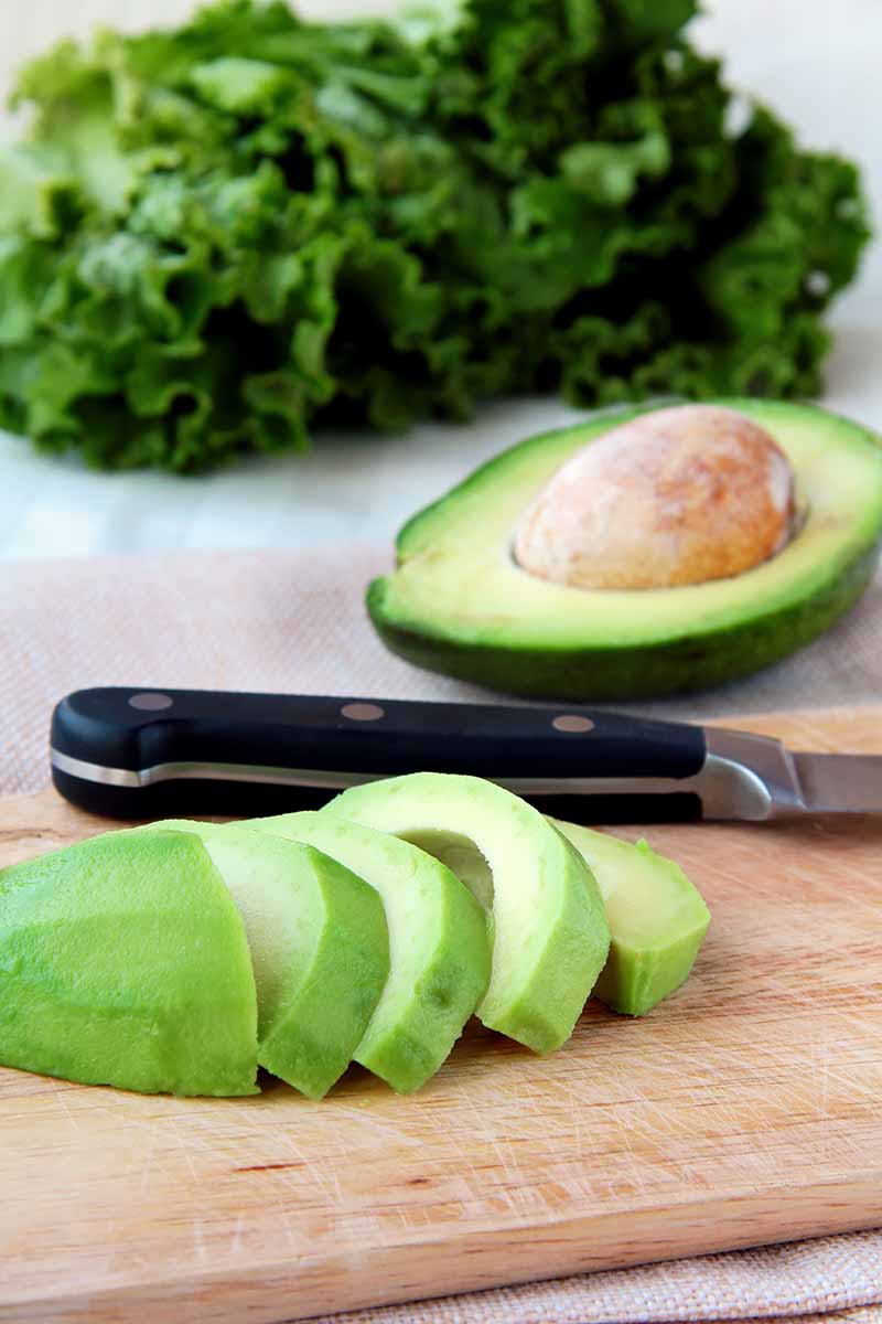 Vertical image of slicing an avocado on a wooden cutting board, with kale in the background.
