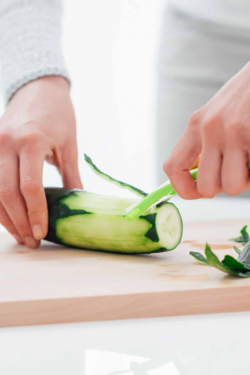 Vertical image of a woman cleaning raw cucumber on a wooden cutting board.