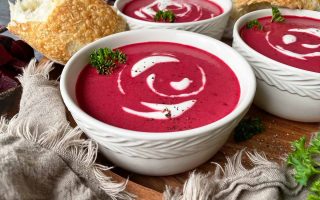 Horizontal image of multiple bowls filled with a bright magenta puree with sour cream swirls and parsley garnish next to bread and more parsley.