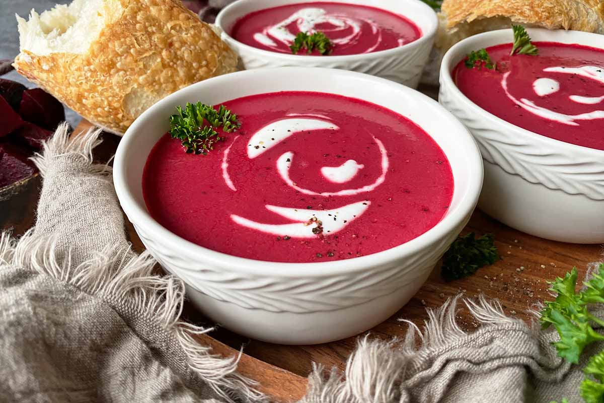 Horizontal image of multiple bowls filled with a bright magenta puree with sour cream swirls and parsley garnish next to bread and more parsley.