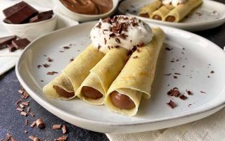 Horizontal image of three rolled thin pancakes with a pudding filling topped with whipped cream.