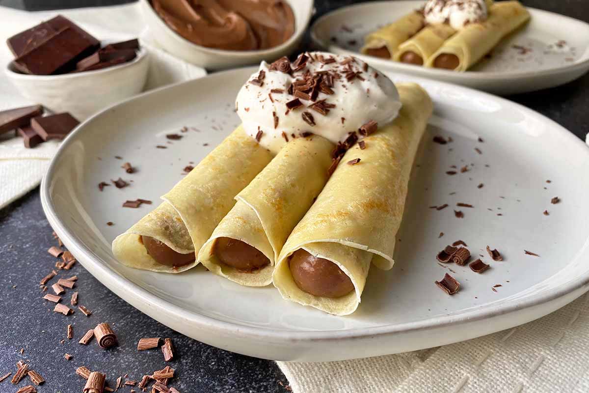 Horizontal image of three rolled thin pancakes with a pudding filling topped with whipped cream.