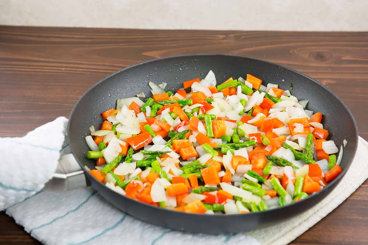 Horizontal image of cooking bell pepper, onion, and asparagus in a skillet with oil.