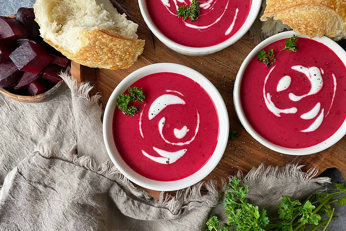 Horizontal top-down image of multiple bowls filled with a bright magenta broth with sour cream swirls and parsley garnish next to bread and more parsley.
