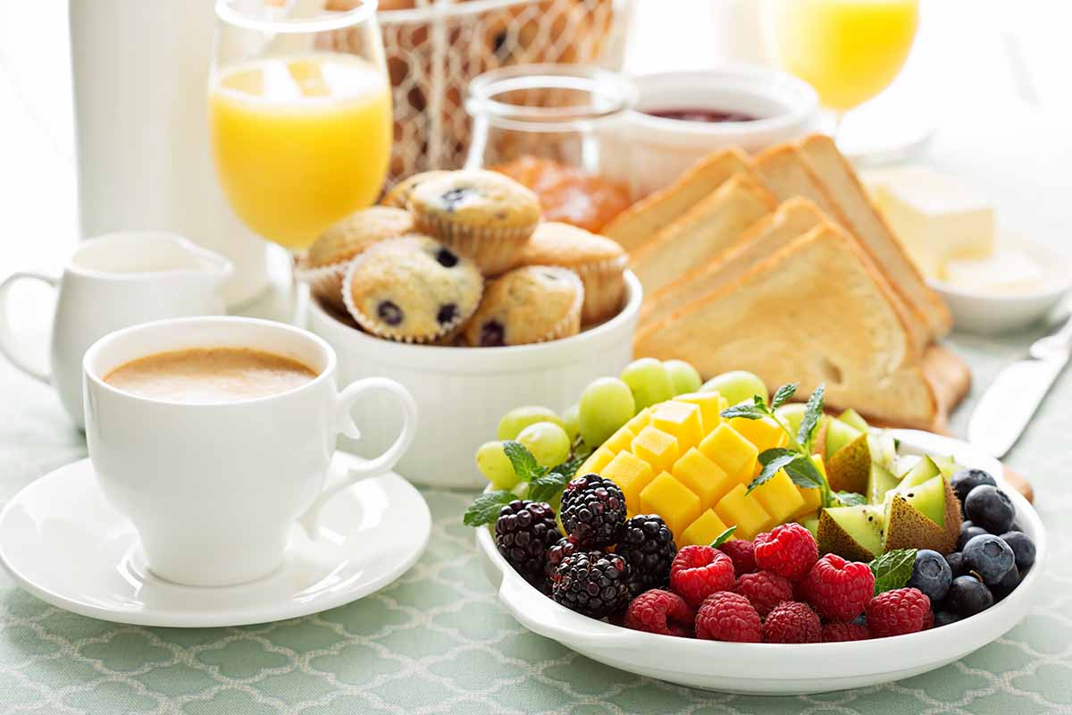 Horizontal image of a table with a bowl of fruit, platter of toast and jam, bowl of muffins, and cups of coffee and orange juice.