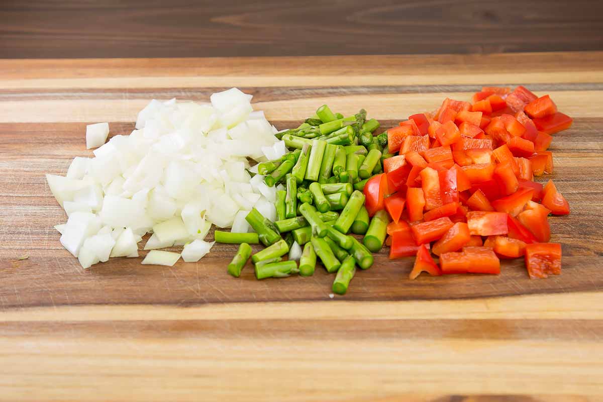 Horizontal image of diced onion, asparagus, and bell pepper on a wooden cutting board.