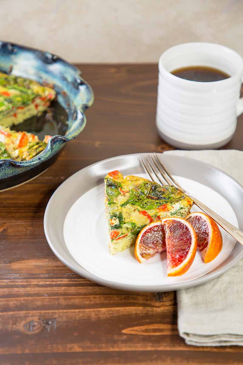 Vertical image of a plate with a wedge of frittata next to orange slices, with a cup of coffee and a napkin.