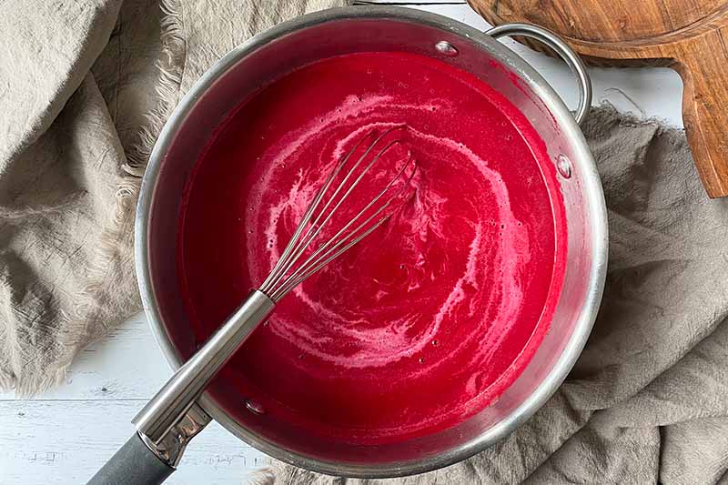 Horizontal image of whisking cream into a bright red stew base in a pan.