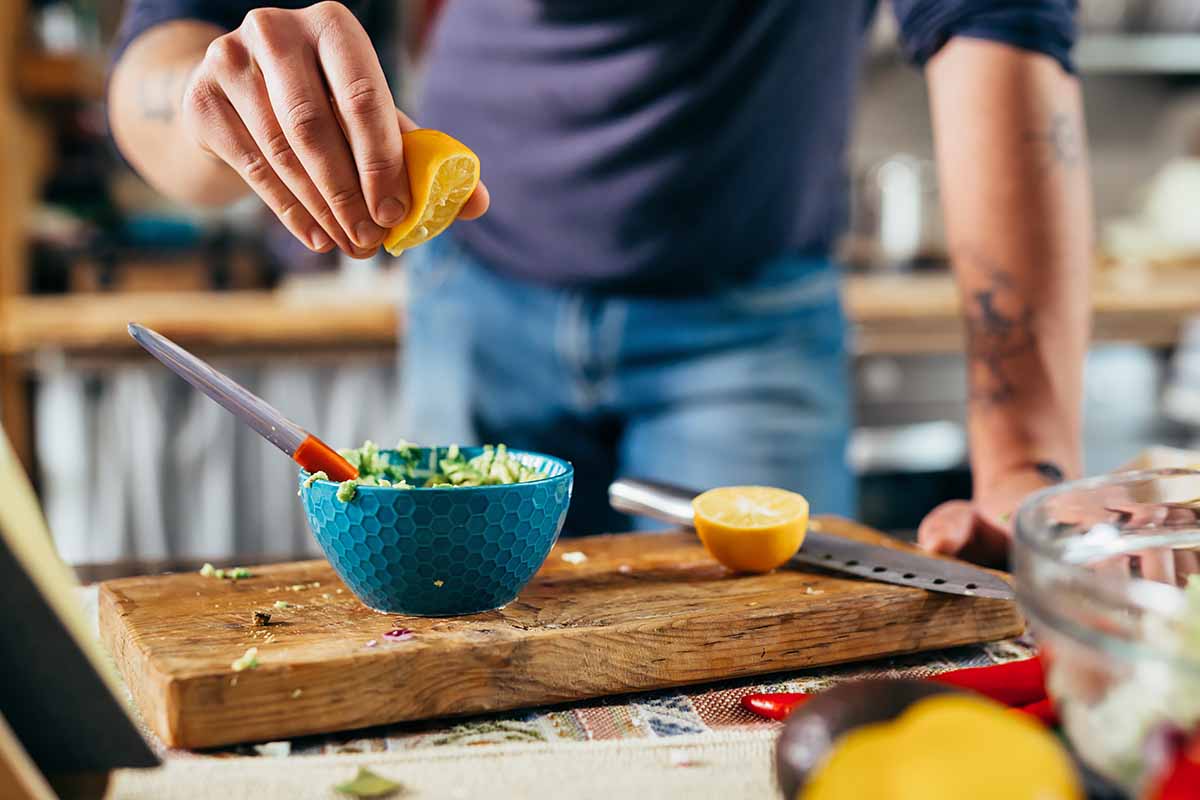 Horizontal image of man squeezing lemon over a dip in a small blue bowl on a wooden cutting board.