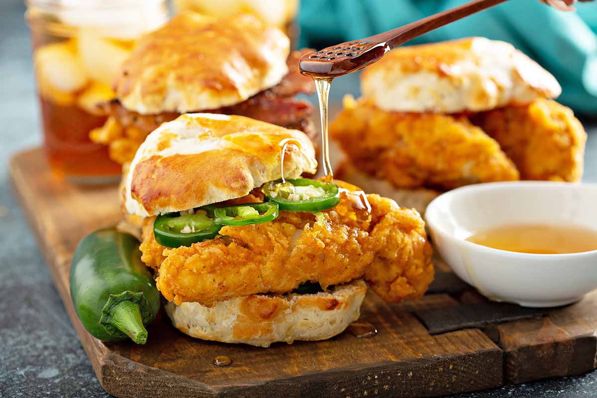 Horizontal image of a breakfast biscuit sandwich with fried chicken and slices of jalapeno, with honey being drizzled over one sandwich.