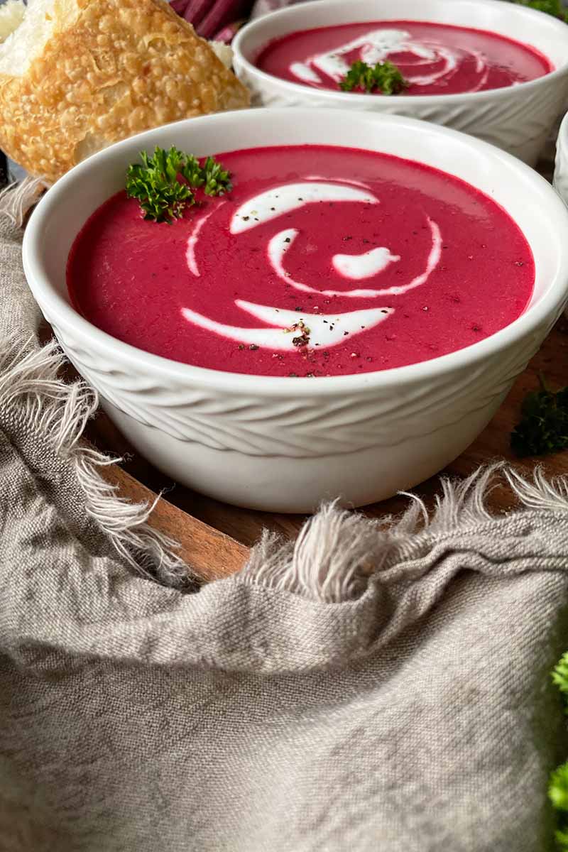 Vertical image of two white bowls filled with a bright magenta puree with sour cream and parsley garnish next to bread and a tan towel.