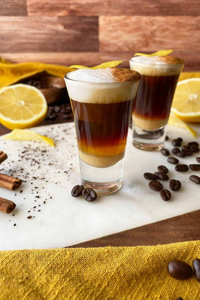 Vertical image of two small shooters filled with a layer espresso drink topped with frothed milk and cinnamon.