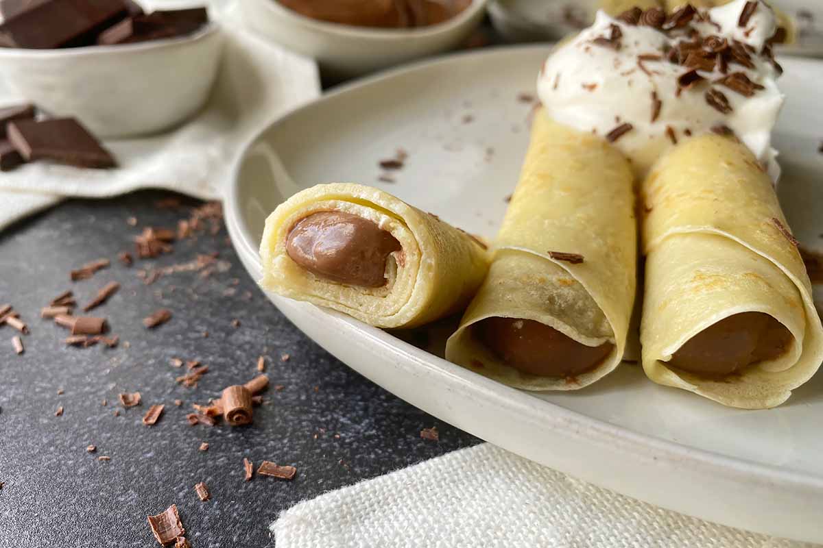 Horizontal close-up image of a halved crepe filled with a chocolate pudding next to other assembled crepes on a white plate.