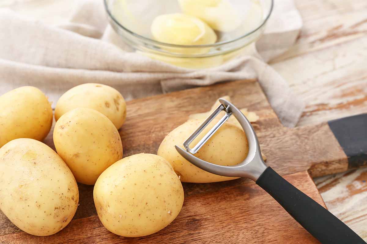 Horizontal image of cleaning raw yellow potatoes on a wooden cutting, with a bowl in the background.