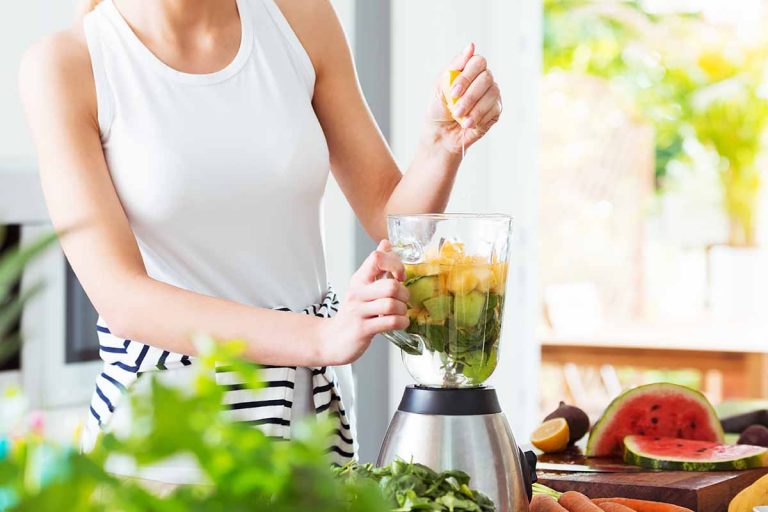Horizontal image of a woman making a fresh shake in the kitchen on a counter next to sliced fresh fruit and vegetables.